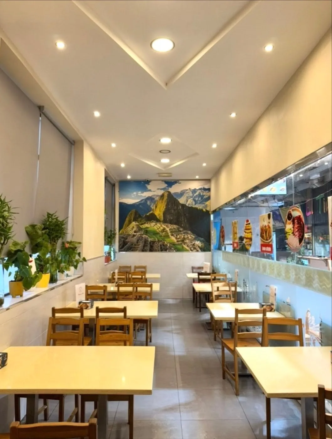 Comedor alargado con mesas de madera clara, plantas en la ventana y un mural de Machu Picchu al fondo